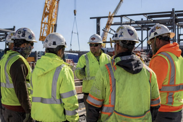 Group of construction workers wearing hard hats and high-visibility jackets gathered around a supervisor during a safety briefing on an active jobsite with cranes and steel structure in the background.